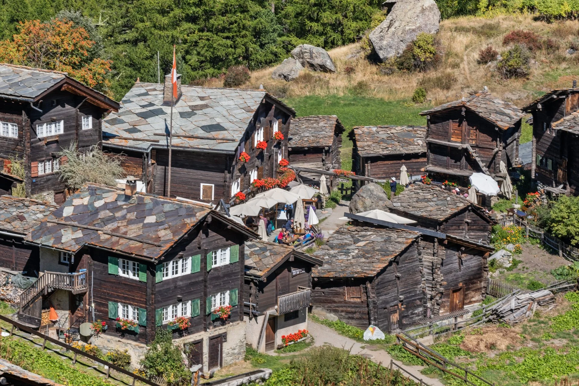 Blick auf traditionelle Holzhäuser in einer alpinen Bergregion mit vielen Menschen, die im Freien sitzen und das schöne Wetter genießen, umgeben von grünen Wäldern und Felsen.