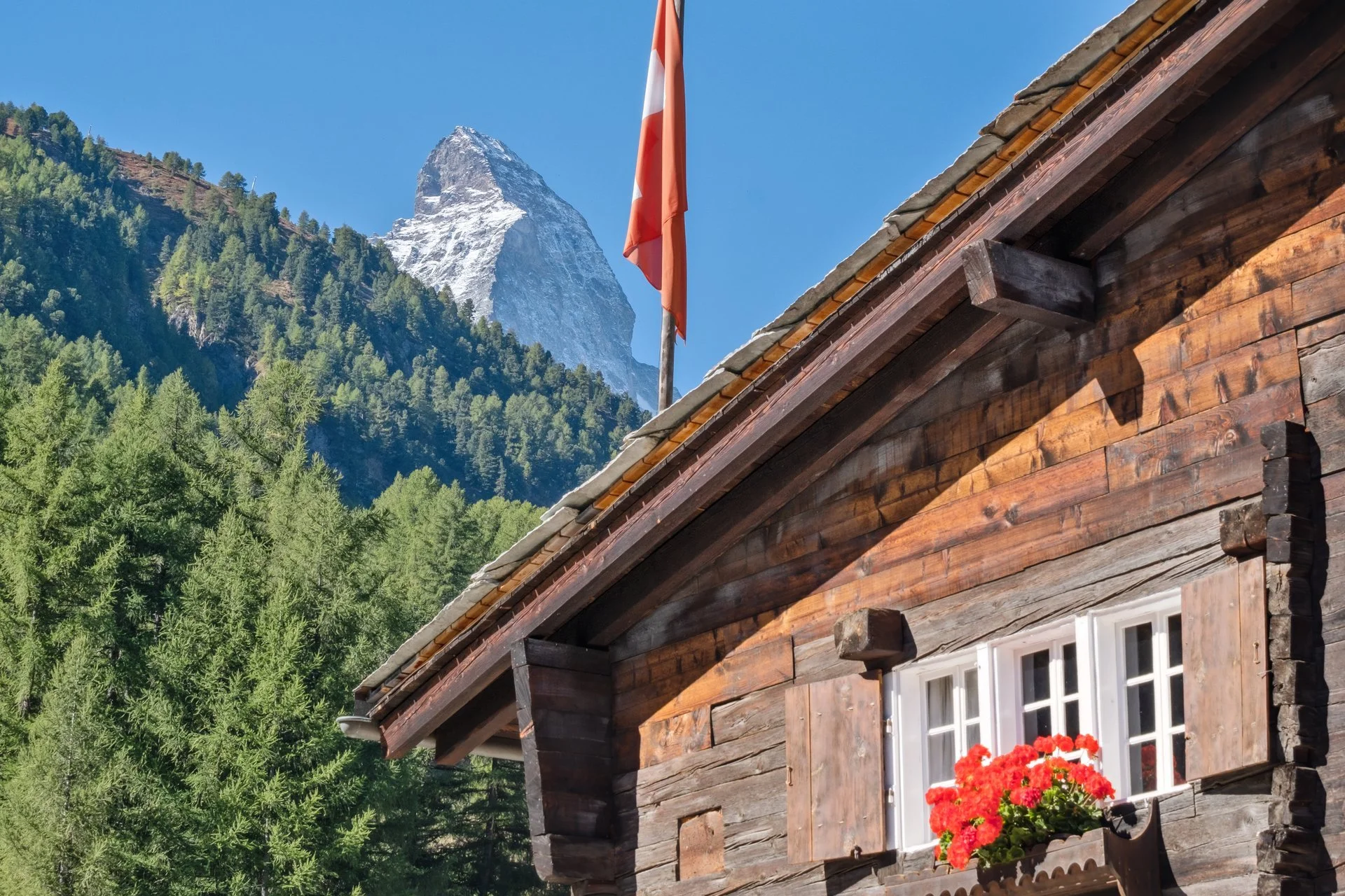 Holzhaus mit weißem Fenster und roten Blumen vor einem Hintergrund aus grünen Bäumen und einem schneebedeckten Berg bei blauem Himmel.