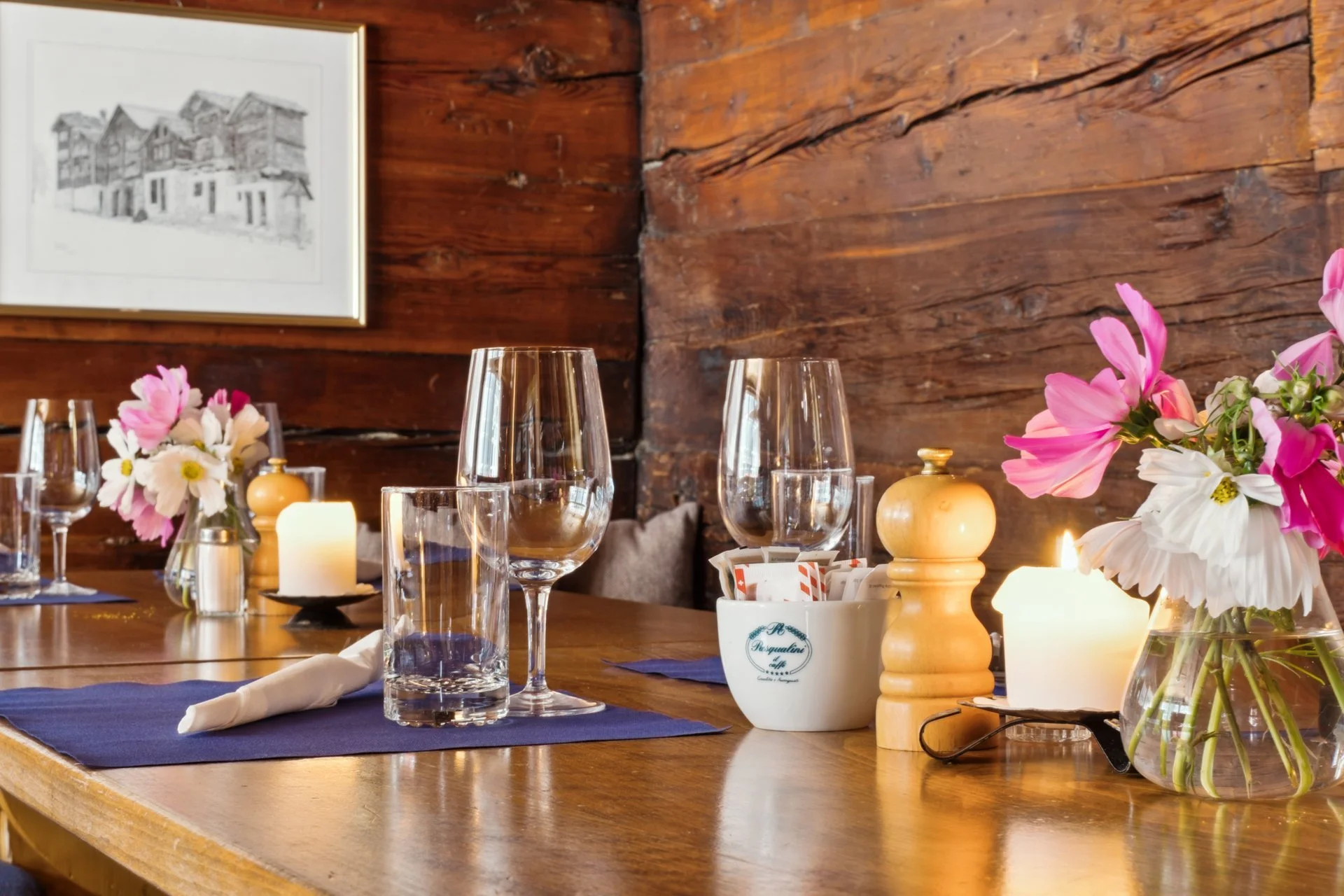 Ein gedeckter Tisch mit Wein- und Wassergläsern, Kerzen, süßem Gebäck, Frühlingsblumen und einem Holz-Hintergrund in einem rustikalen Raum.
