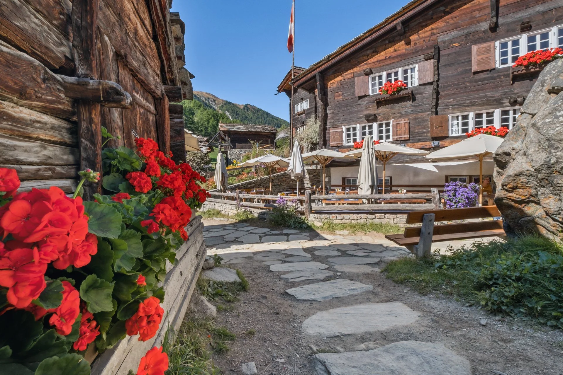 Ein idyllischer Bergort mit alten Holzgebäuden, einer Terrasse mit Stühlen und Sonnenschirmen, sowie bunten Blumen im Vordergrund und grünen Bergen im Hintergrund.