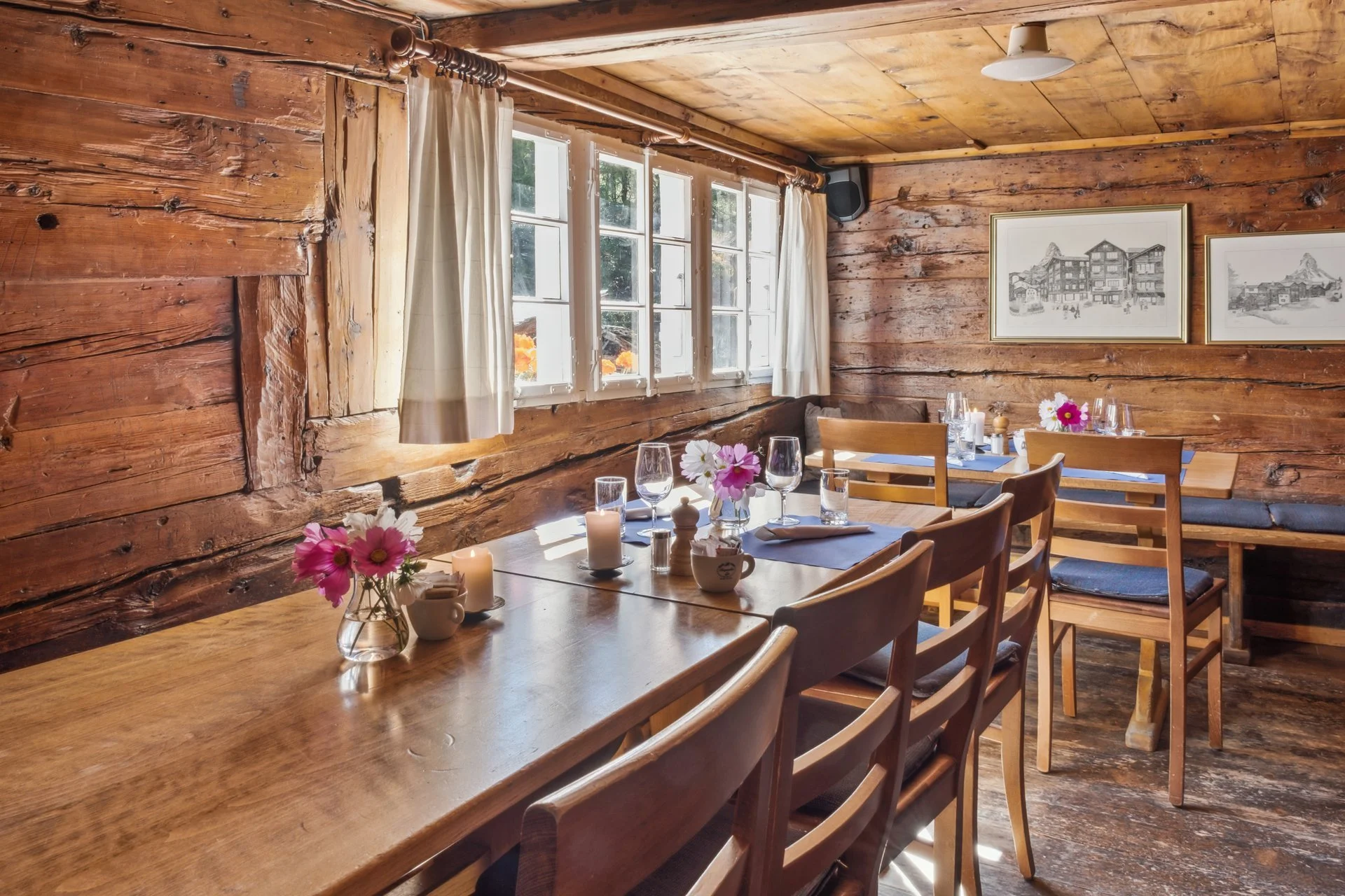 Ein rustikaler Speiseraum mit Holzwänden, großen Fenstern, gedecktem Tisch mit Blumen, Kerzen und Geschirr, an den Wänden hängen Schwarz-Weiß-Bilder.