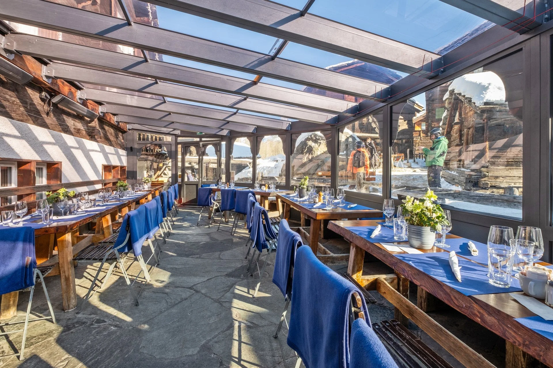 Innenaufnahme eines gemütlichen Restaurant mit Glasdach, gedecktem Tisch, blauen Stuhlhussen, Wein- und Wassergläsern, und Winterlandschaft im Hintergrund.