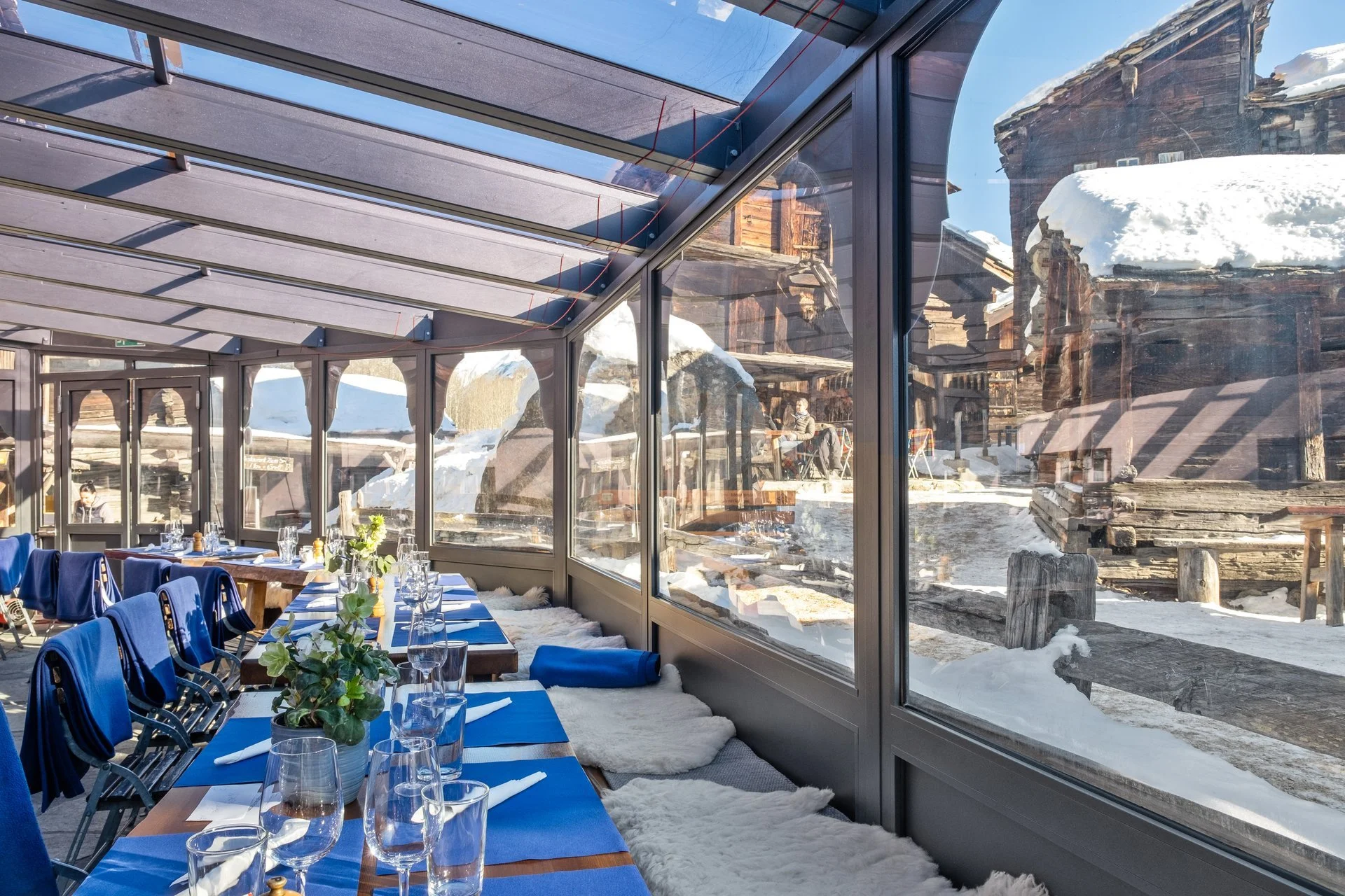 Innenraum eines Restaurants mit großen Glasfenstern, Tischen mit blauen Tischdecken, Gläsern und Blumen, Blick nach draußen auf schneebedeckte Holzgebäude in einer winterlichen Umgebung.