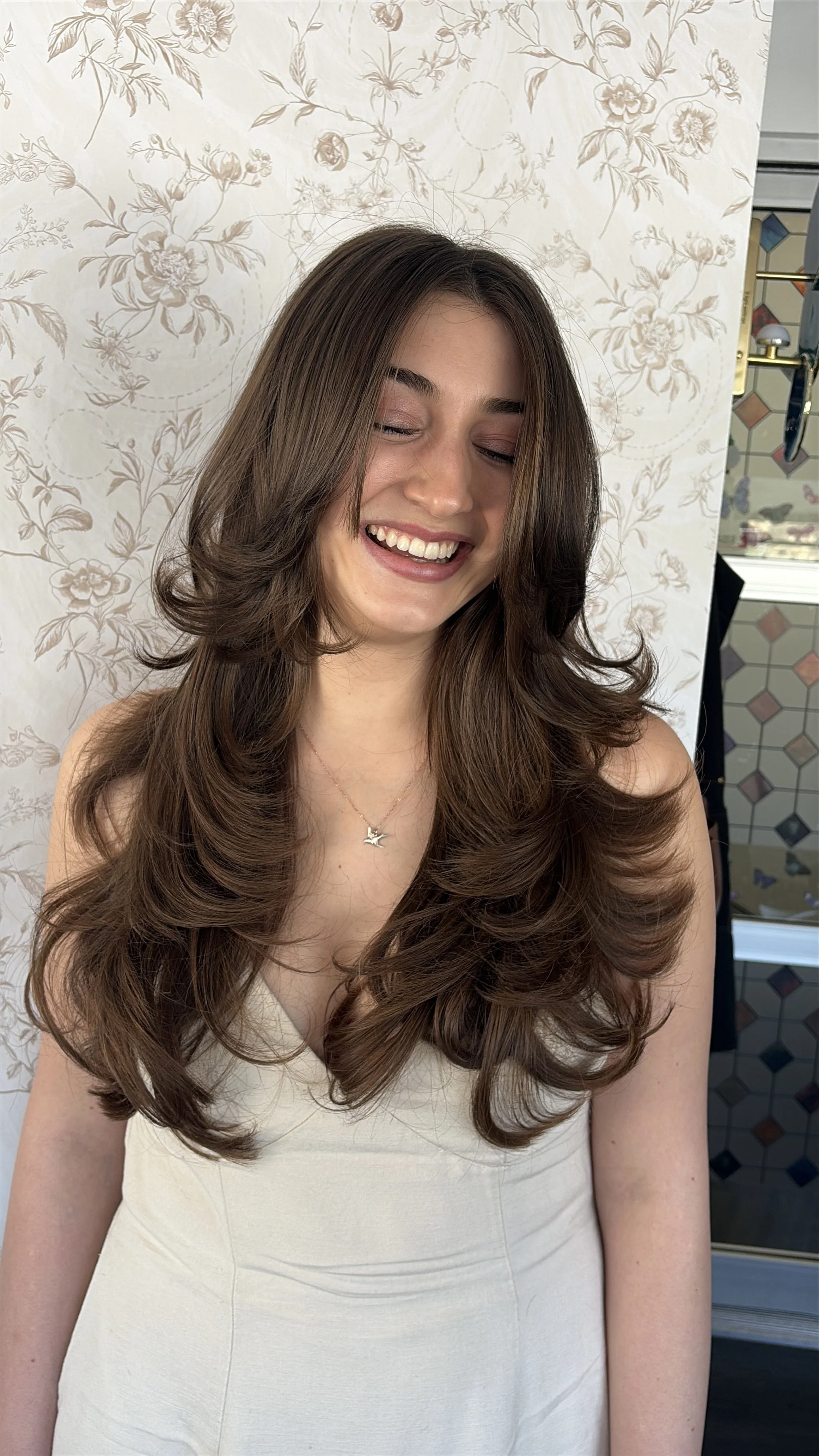 A young woman with long, wavy brown hair and a big smile, wearing a white dress and a silver bird necklace, standing in front of a floral wallpaper background.