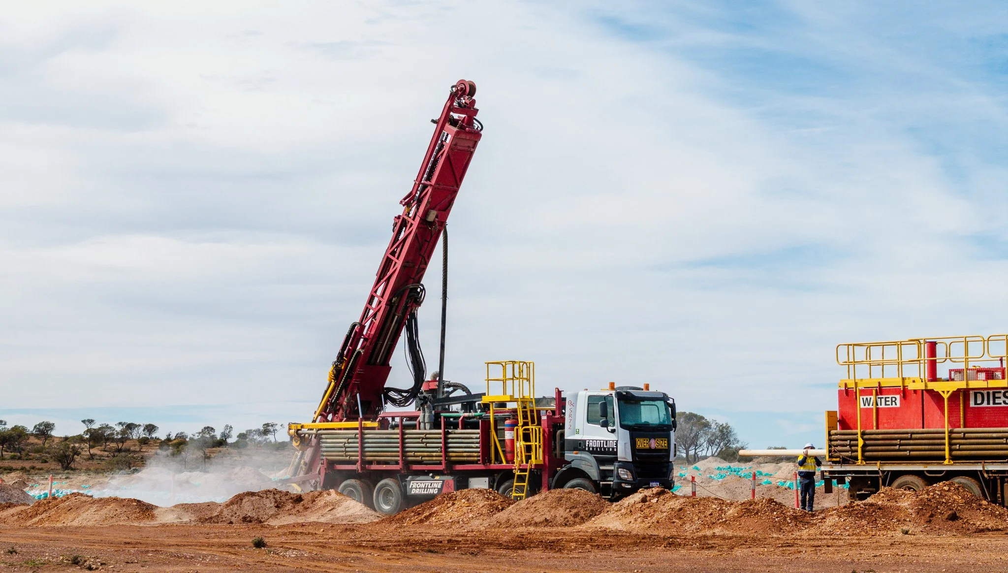 A large red Frontline Drilling rig mounted on a truck, operating on a mining exploration site with a clear sky and some workers nearby.