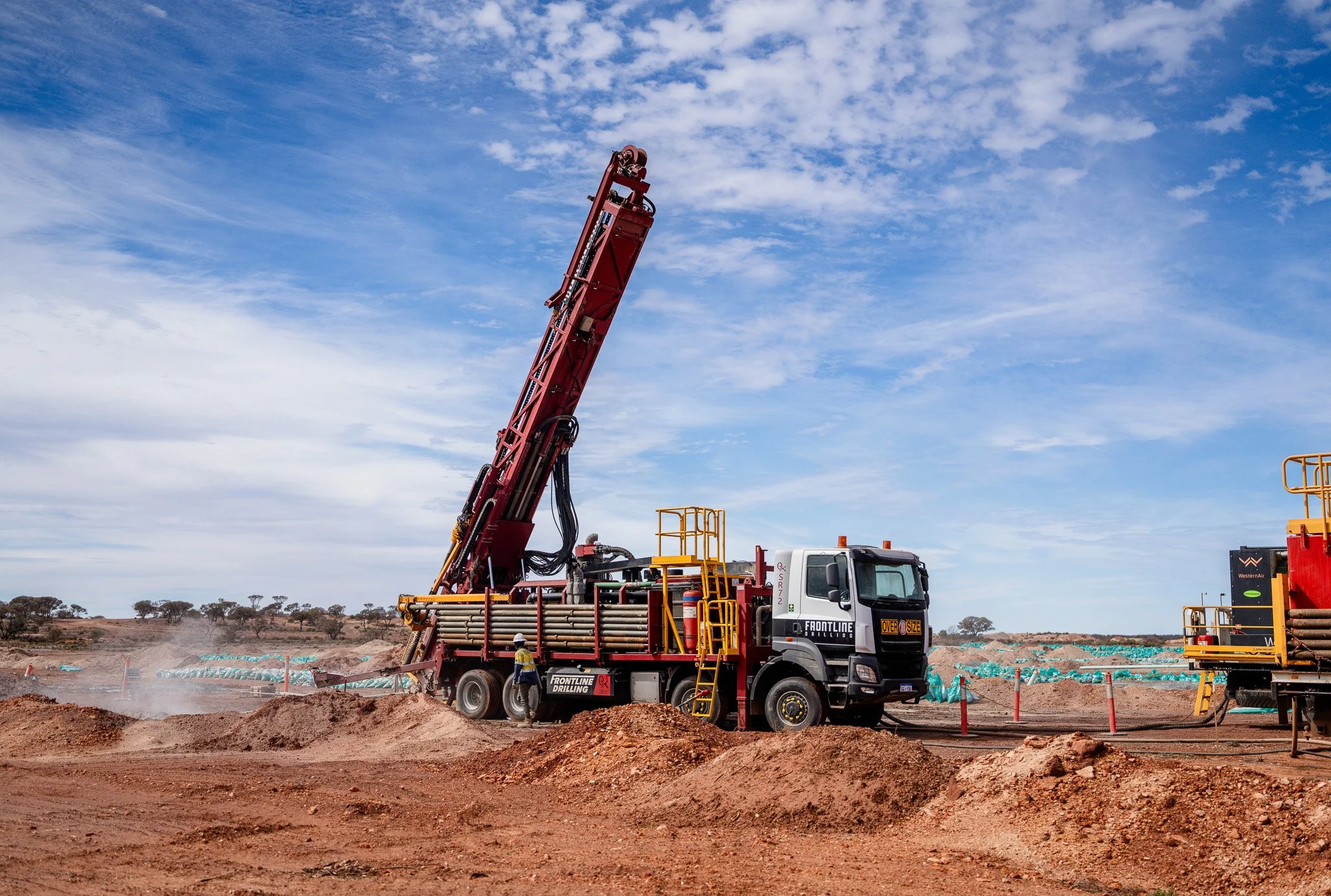 A large red drilling machine mounted on a truck at an outdoor construction site, with soil and construction materials in the foreground and a partly cloudy blue sky in the background.