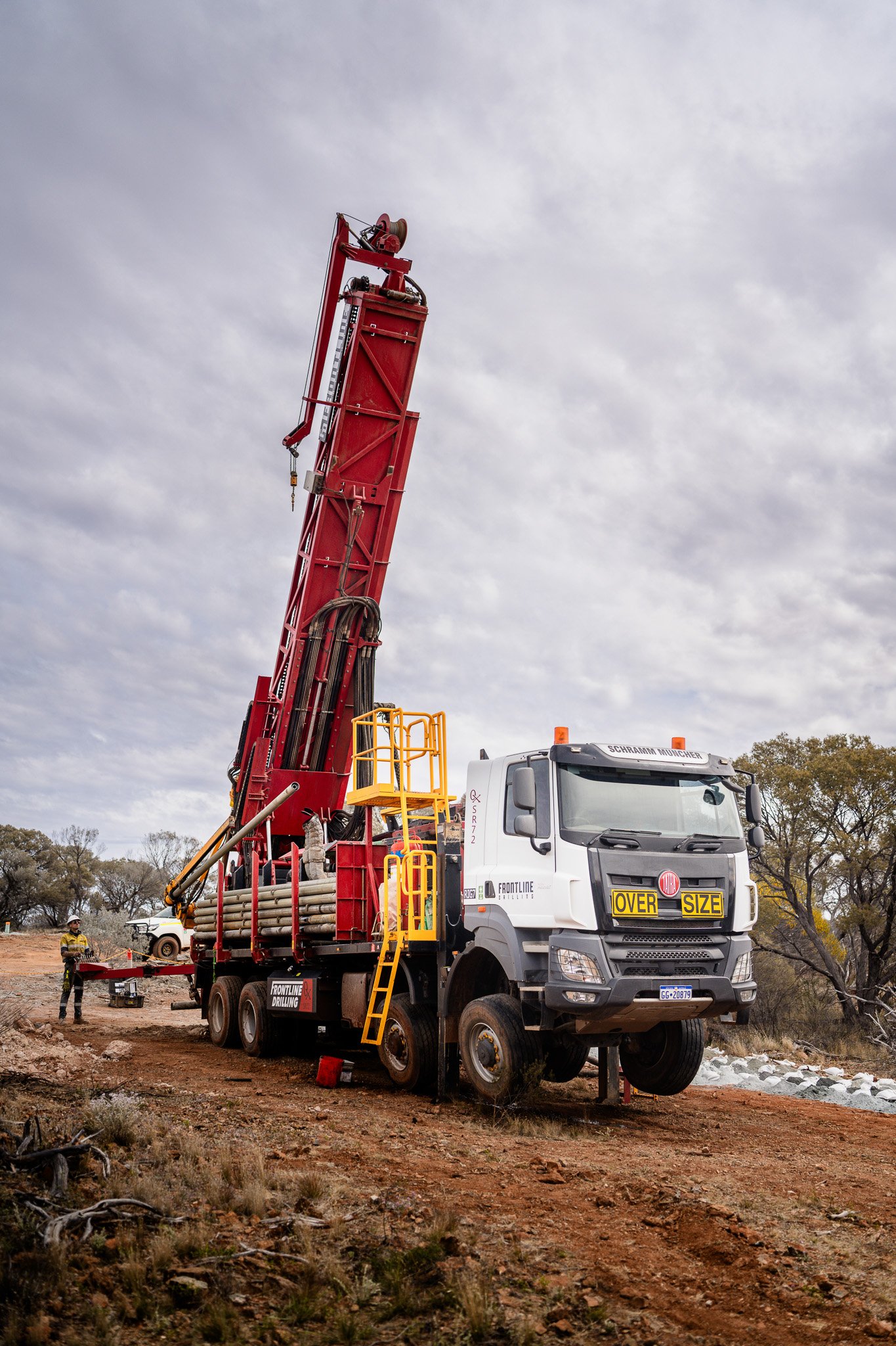 A large red Frontline Drilling rig on a mining explortation site with a cloudy sky in the background. There are workers and trees visible around the site.