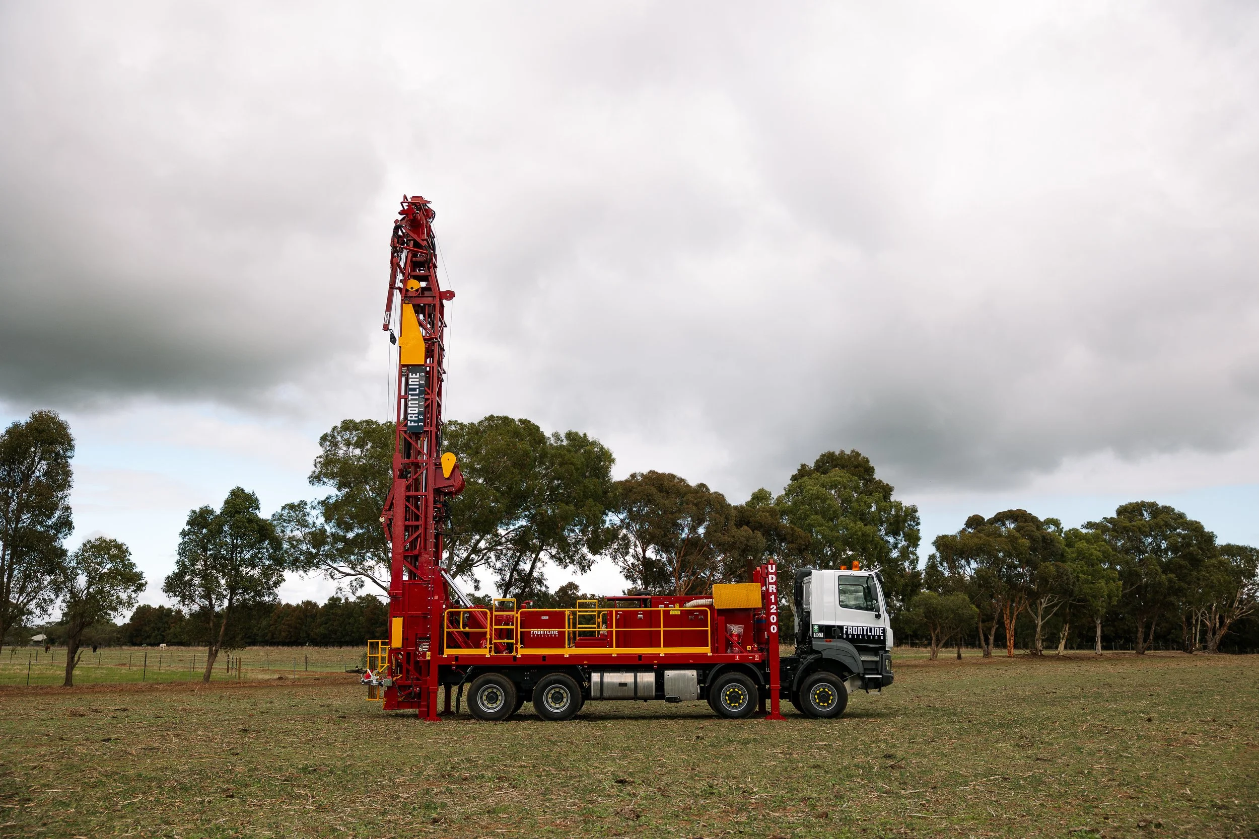 A large red Frontline Drilling rig mounted on a truck in a grassy field under a cloudy sky, with trees in the background.