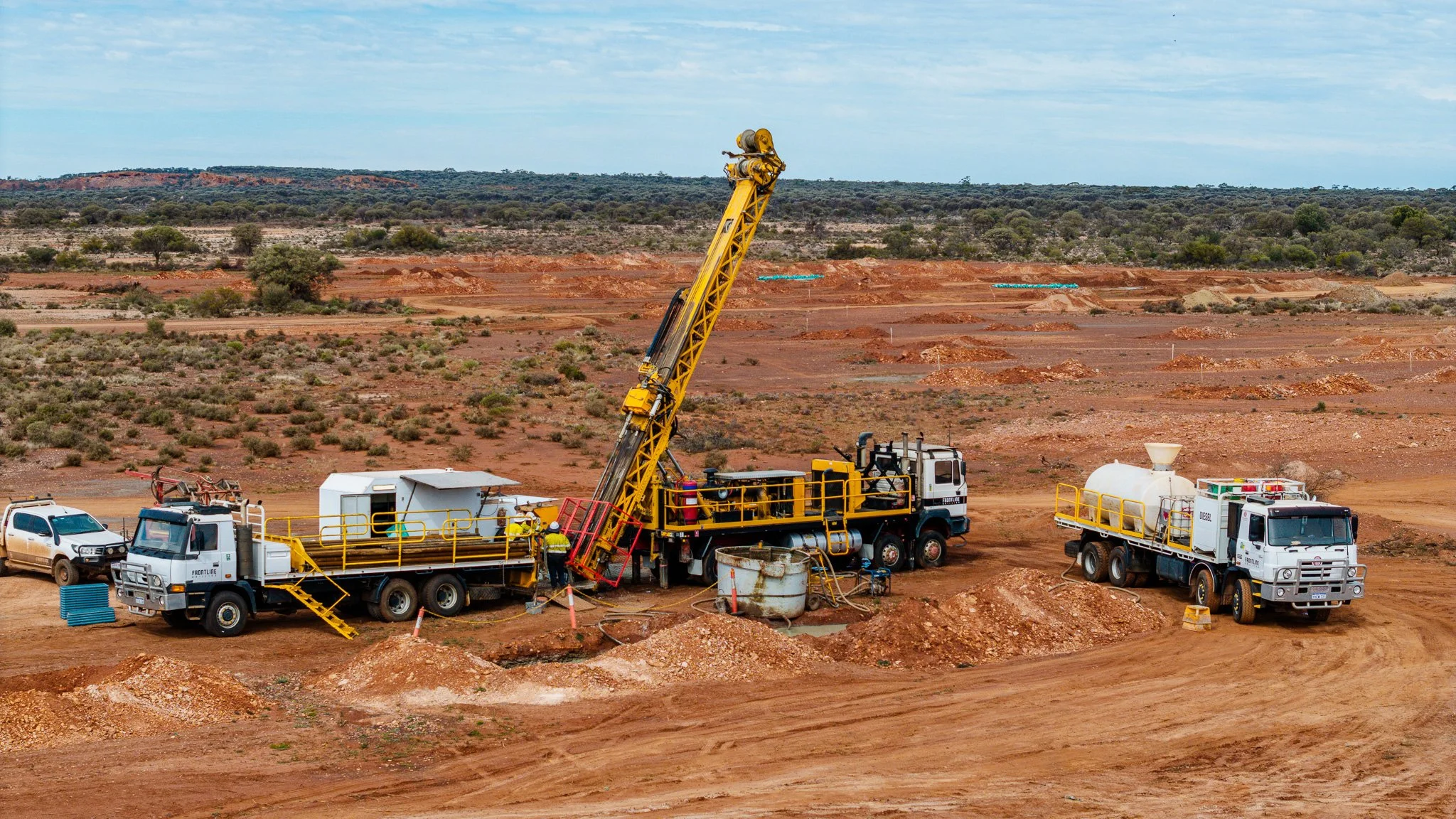 Mine site with large yellow drill rig, multiple trucks, and workers on red dirt landscape, with sparse vegetation and distant hills under a partly cloudy sky.