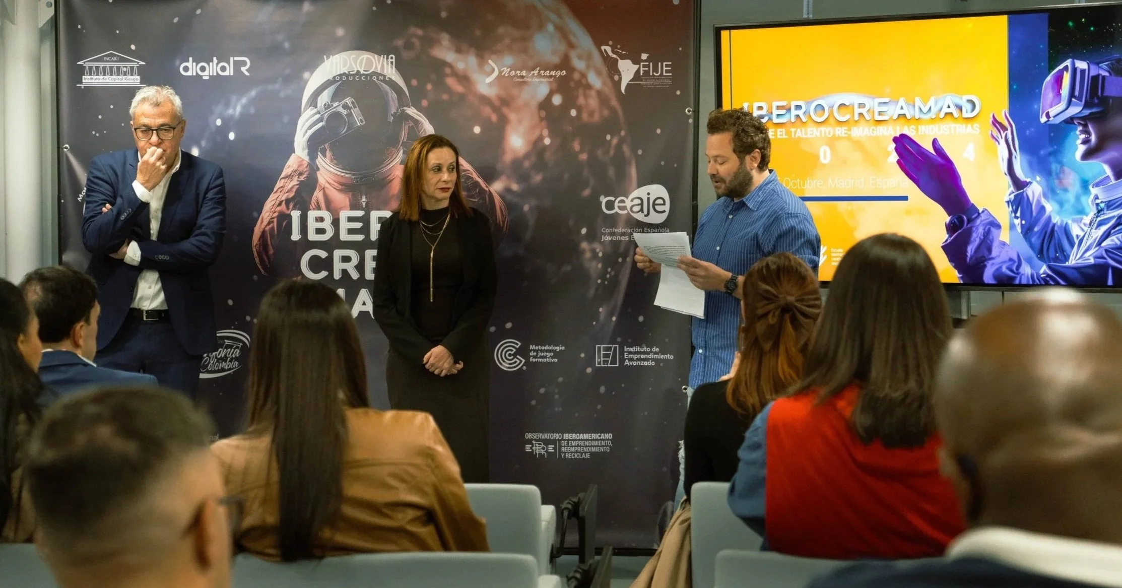 Una conferencia con varias personas en un auditorio, con un fondo con texto y logotipos, y una pantalla grande que dice 'HUBEROCREAMAD' y 'REIMAGINA EL TALENTO RE-IMAGINA LAS INDUSTRIAS'.