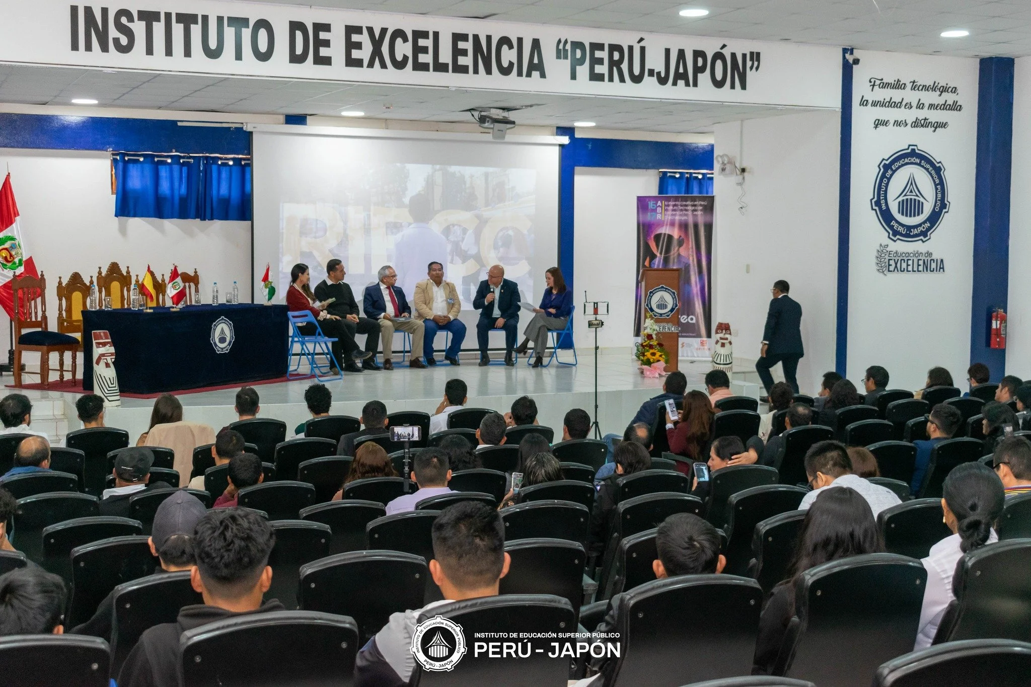 Una audiencia en un auditorio con varias personas sentadas en sillas negras y una mesa al frente con banderas de Perú, España, Palestina, y un logotipo de un instituto. En el escenario, cinco personas dialogan, una de ellas con micrófono. A la derecha, una persona camina. Hay un cartel grande arriba con el texto 'INSTITUTO DE EXCELENCIA "PERÚ-JAPÓN"' y un cartel a la derecha del logo del instituto que dice 'Familia tecnológica, la unidad es la medalla que nos distingue'.