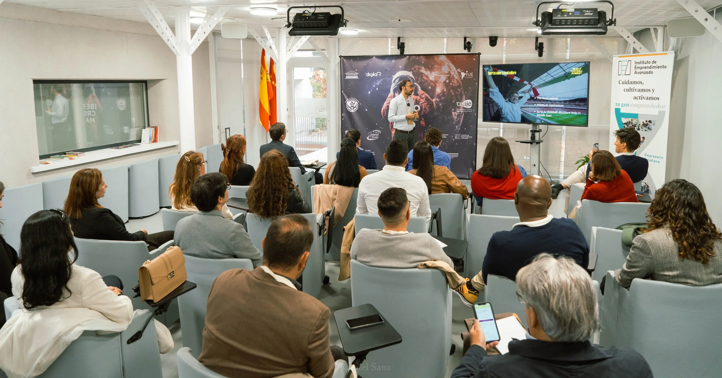Una conferencia con varias personas asistiendo, un presentador en el escenario con micrófono, pantallas y banners de fondo, bandera de España a la izquierda.