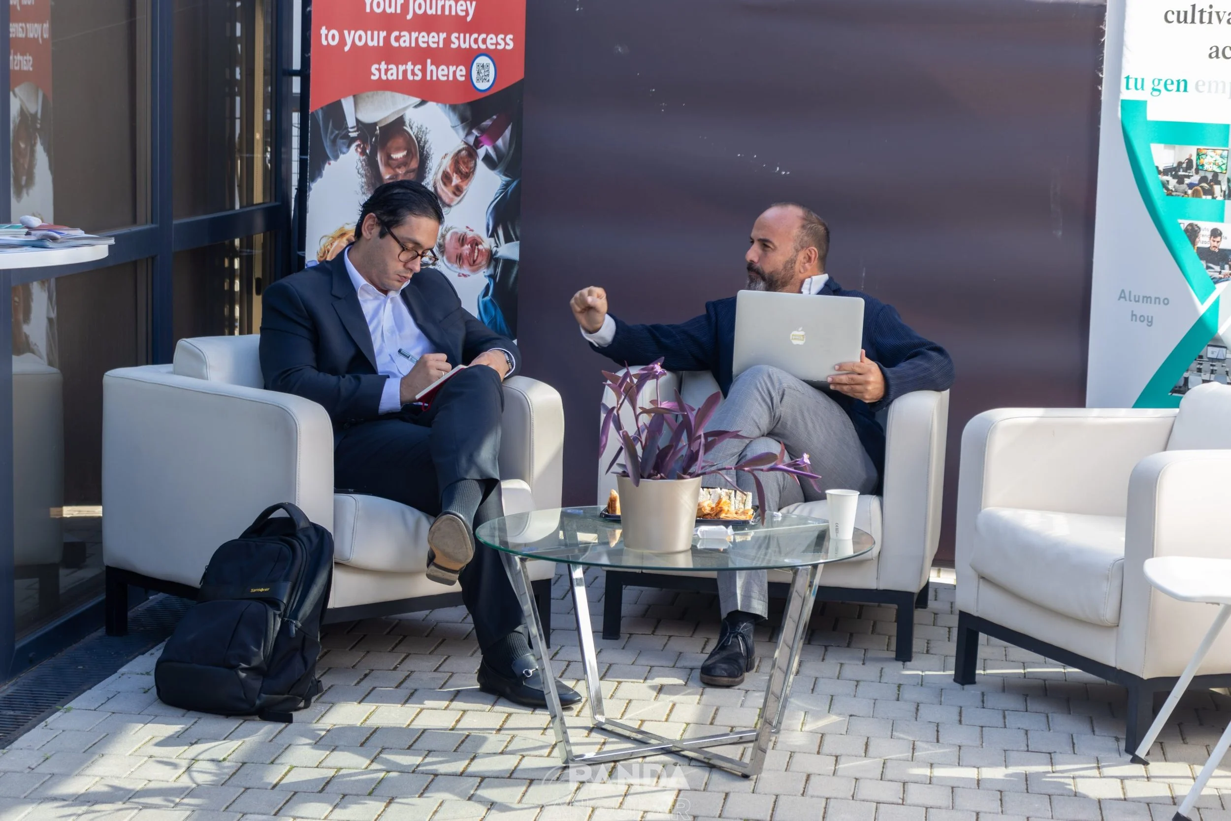 Dos hombres sentados en sillones blancos conversando en un espacio exterior, con una mesa de vidrio frente a ellos que tiene comida y una planta en jarrón. Uno de ellos usa una laptop y el otro un cuaderno, y hay un cartel con texto en inglés y fotos en el fondo.