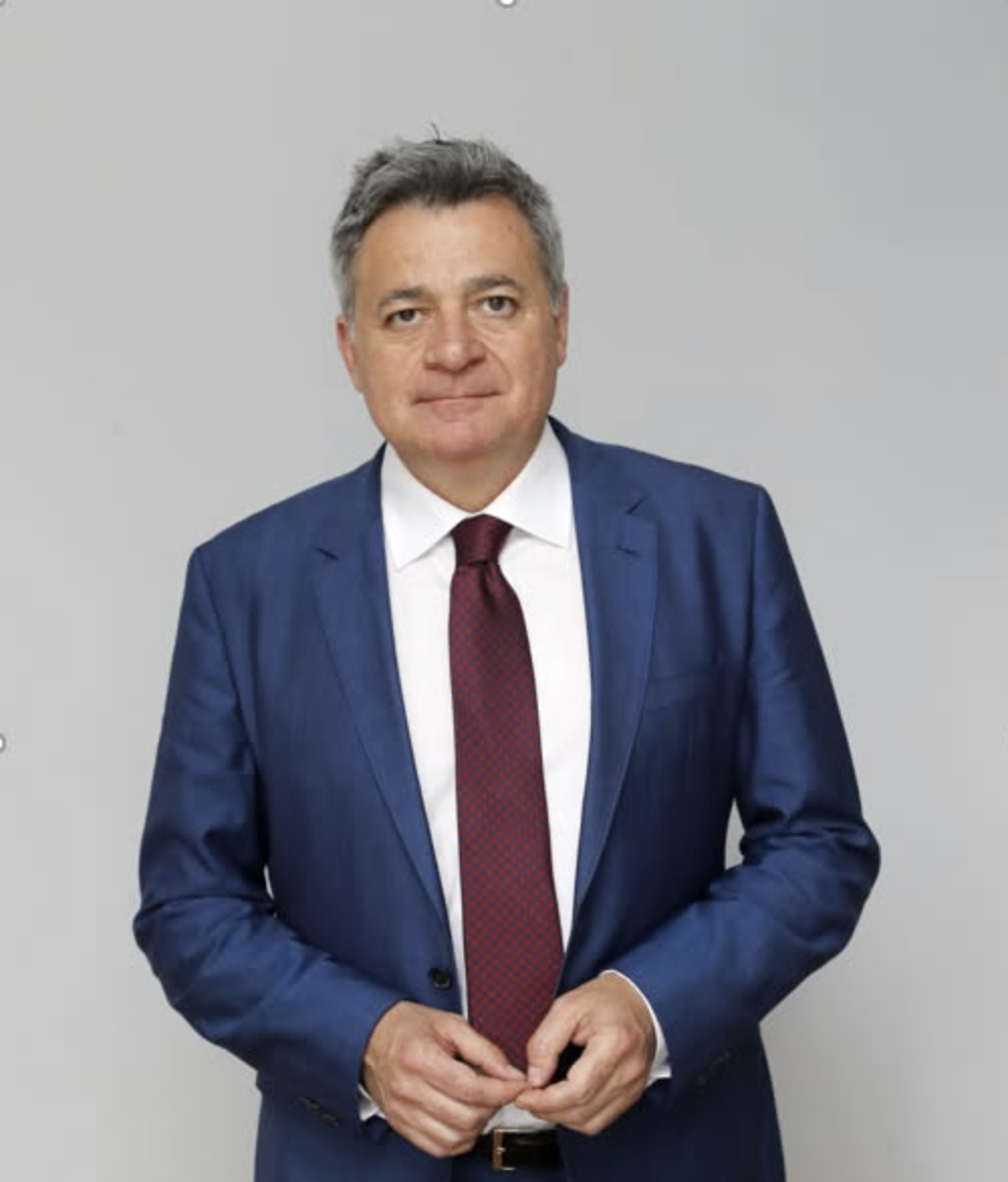 Hombre con cabello gris, vestido con saco azul y corbata roja, frente a un fondo gris claro.