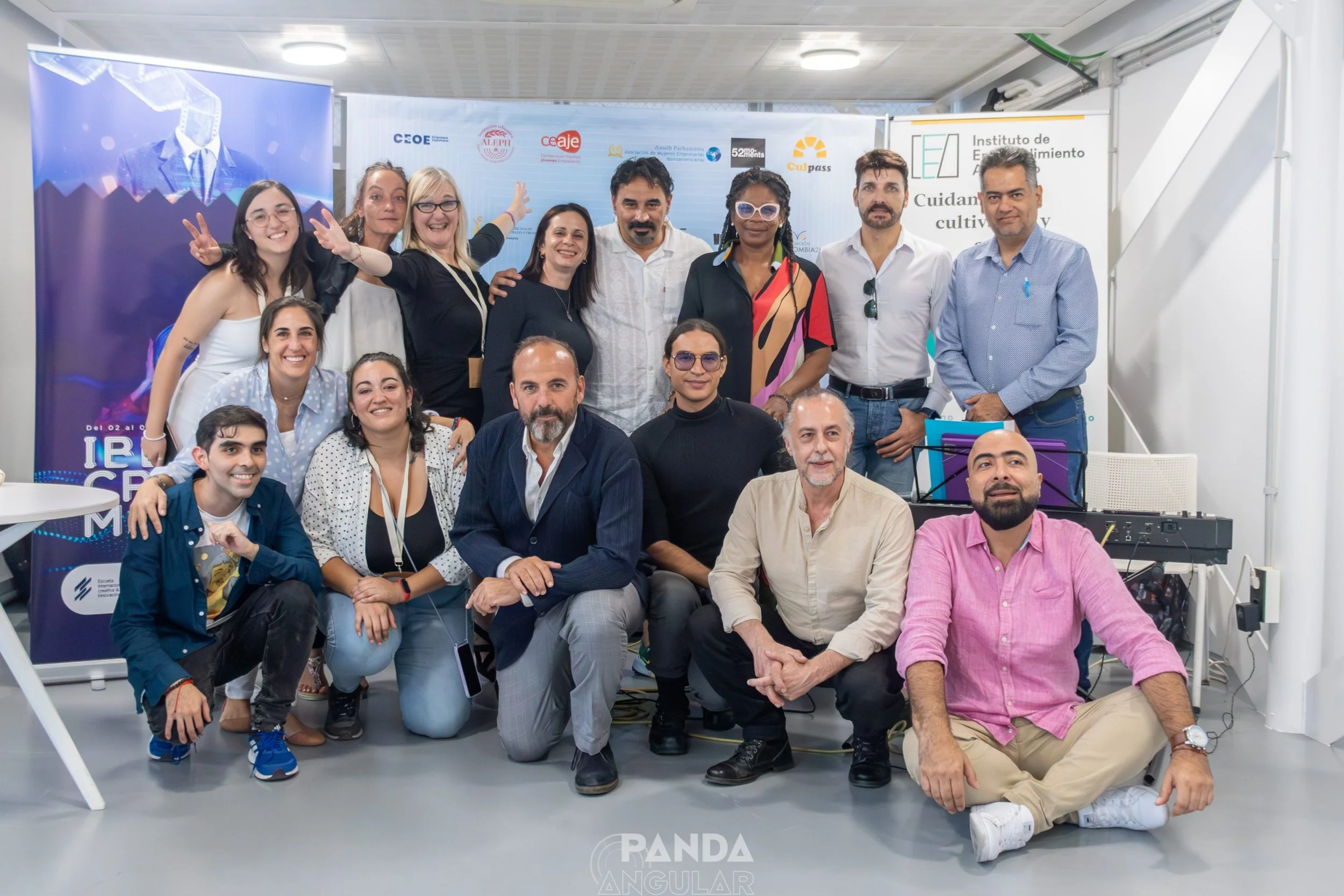 Grupo de personas posando en un evento, algunos son de pie y otros están sentados, en segundo plano hay un banner y una pantalla con gráficos y logotipos relacionados con la innovación y el emprendimiento.