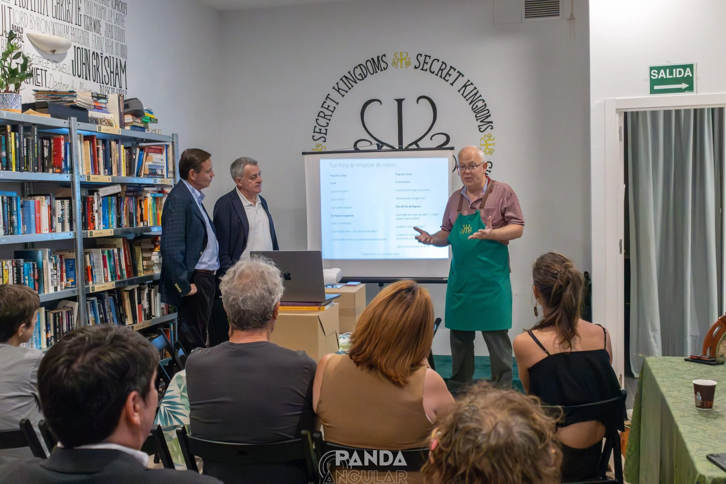 Una sala de conferencias con varias personas de pie y sentadas, un hombre con delantal verde explica frente a una pantalla y varios libros en las estanterías. Personas escuchando la charla, fondo con logotipo de 'Secret Kingdoms' en la pared.