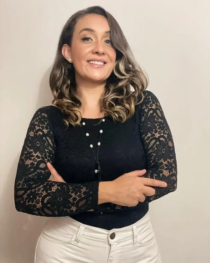 Mujer de cabello rizado y rubio claro, con blusa negra de encaje en las mangas, caminando frente a una pared plana.