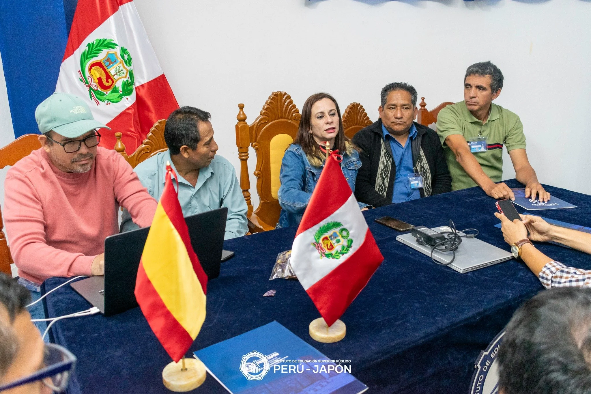 Reunión oficial con cinco personas sentadas en una mesa con banderas de Perú y Japón, en un entorno formal con un fondo de pared blanca y un escudo del Instituto de Educación Superior-PERÚ-JAPÓN en la mesa.