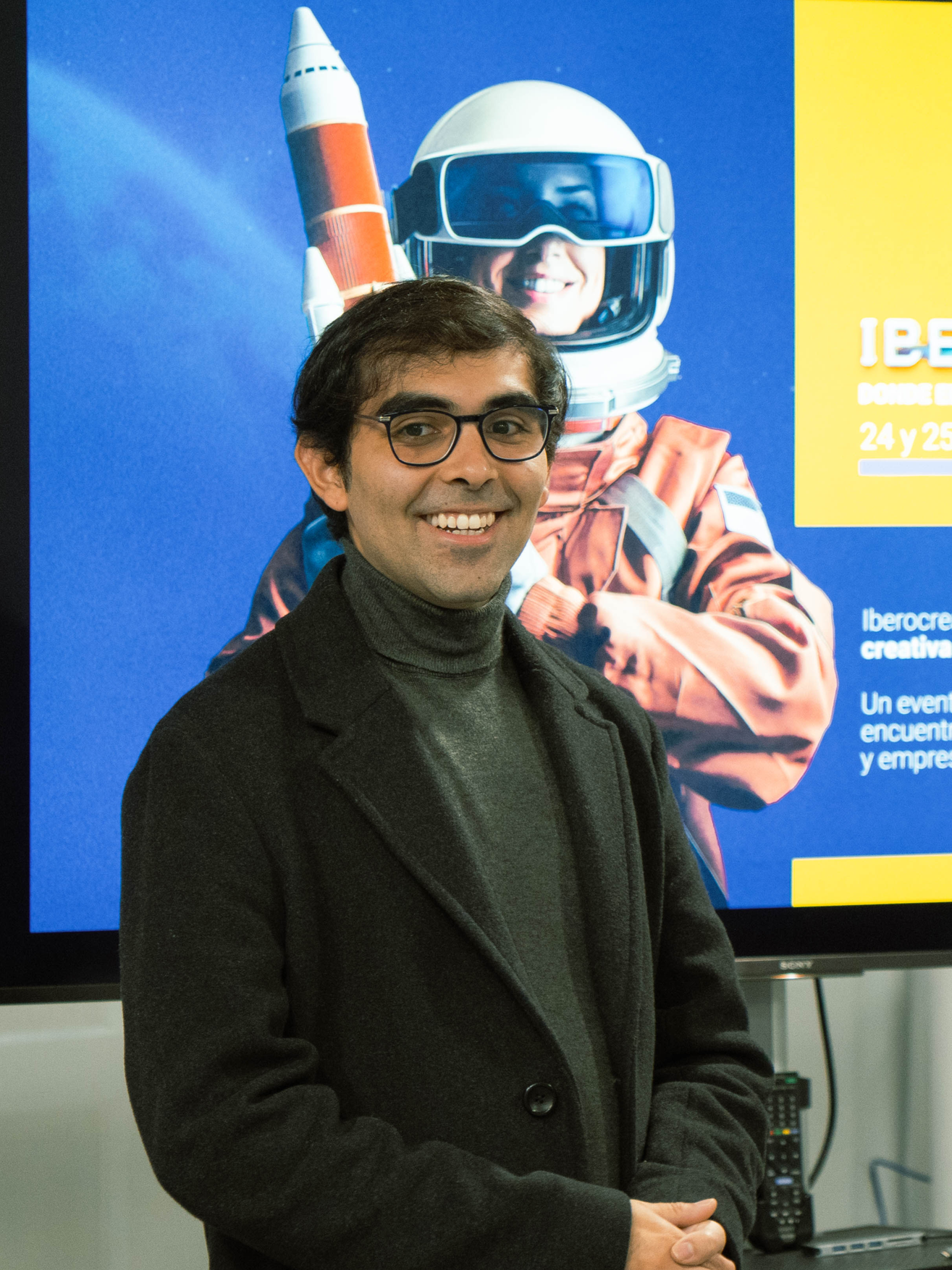 Hombre con gafas y abrigo negro sonriendo frente a un cartel con un astronauta y un cohete.