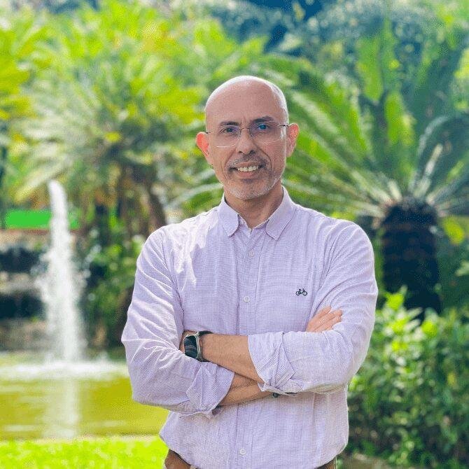 Hombre con gafas, camisa blanca, en un entorno natural con árboles, fuente y vegetación.