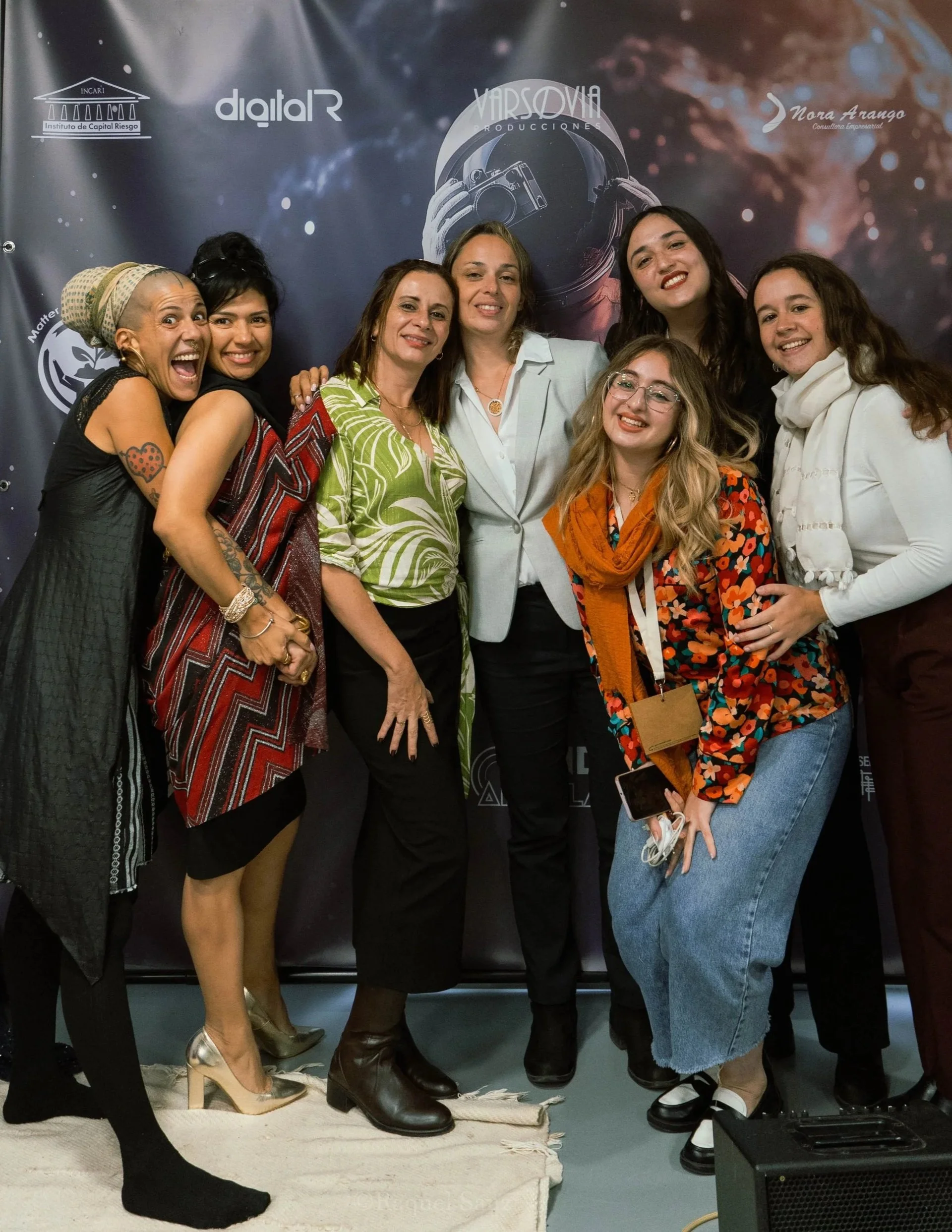 Grupo de mujeres sonriendo y posando juntas en un evento, con un fondo con logos y palabras relacionadas con producción y creatividad.
