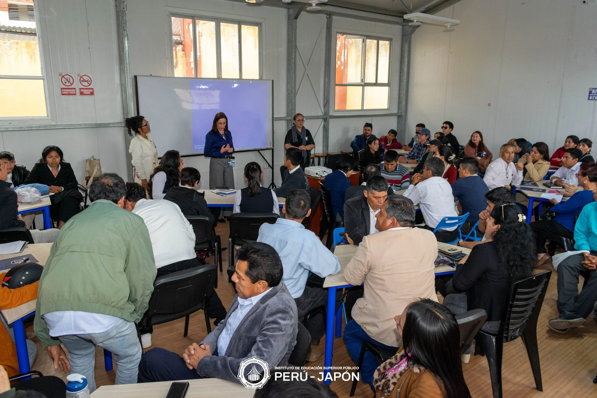 Una sala de clases con muchas personas sentadas en mesas, una profesora en el frente, una pantalla gigante, y a la pared hay un cartel que dice 'Instituto de Educación Superior Público Perú - Japón'.