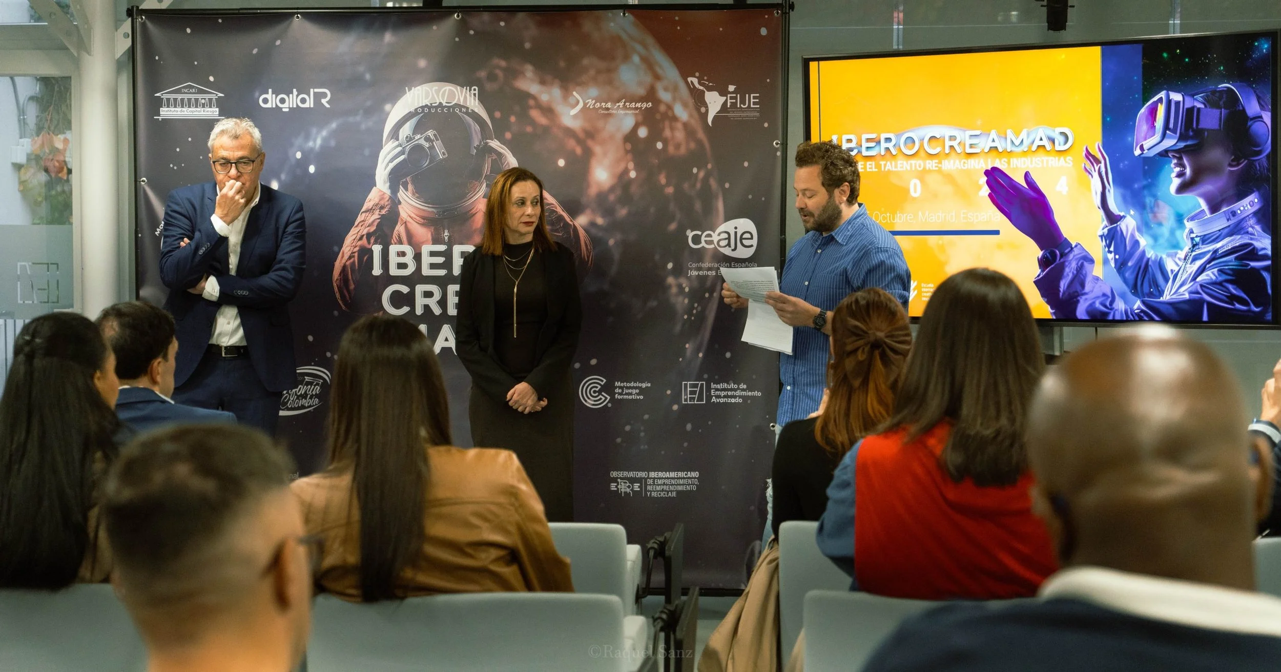 Personas en una conferencia con un fondo que dice "IBEROCREMAD" y una pantalla con una imagen de una mujer con realidad virtual, en un entorno académico o profesional en Madrid, España.