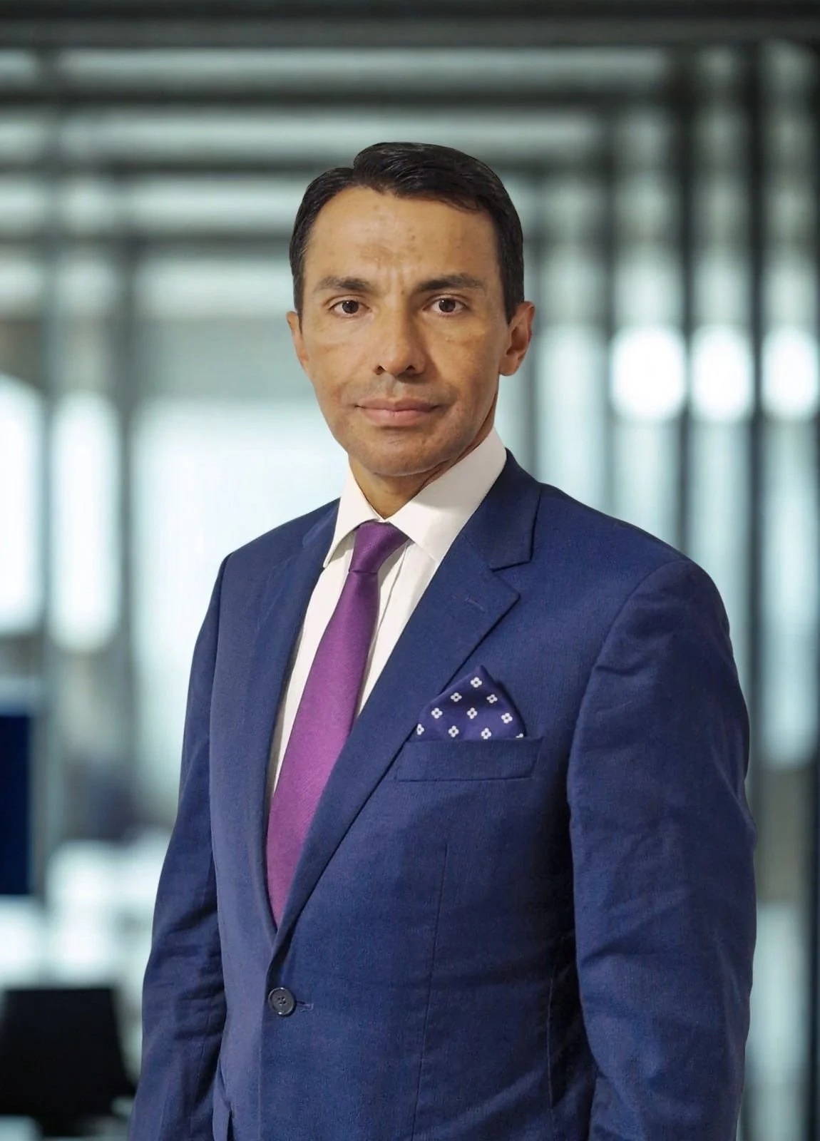 Hombre con barba, complexión delgada, peinado corto, vestido con saco azul, camisa blanca, corbata magenta y pañuelo de bolsillo azul con puntos blancos, en un entorno de oficina moderna con fondo borroso de ventanas grandes.
