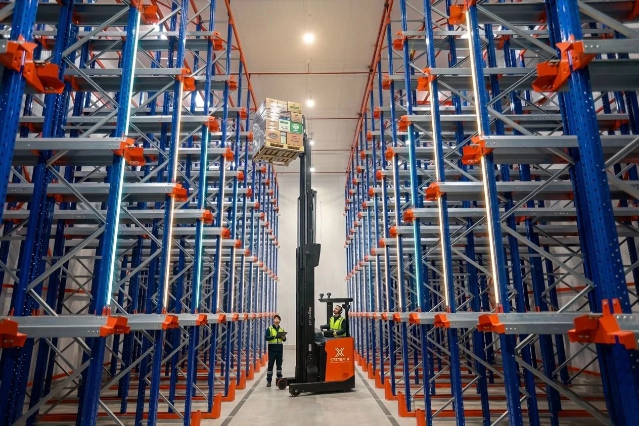Warehouse storage system with tall blue metal shelves, a forklift lifting a pallet of boxes, and two workers in safety vests.