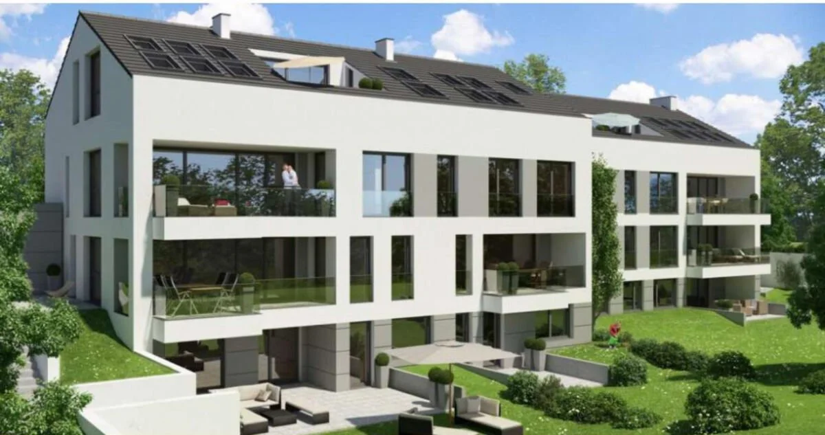 Modernes Mehrfamilienhaus mit weißen Wänden, großen Fenstern, Balkonen und Solarmodulen auf dem Dach, umgeben von grüner Natur.