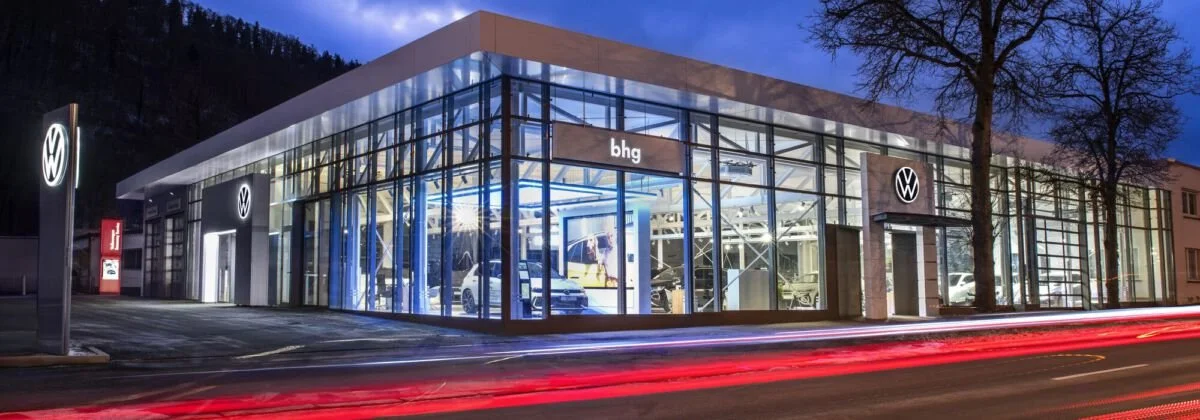 Moderne Volkswagen-Autohändler mit Glasfassade bei Nacht, beleuchtete VW-Logos, einige Fahrzeuge im Showroom sichtbar.