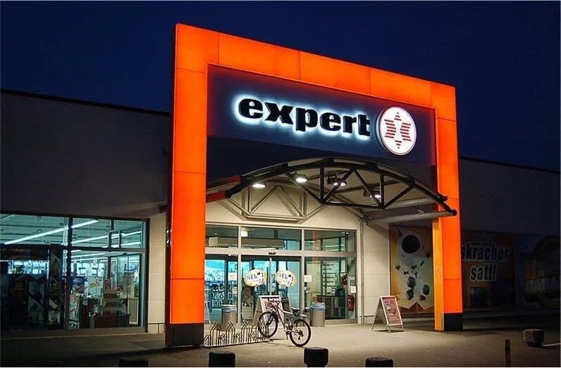 Außenansicht eines Geschäfts mit leuchtendem Schild "expert" bei Nacht, mit Fahrrad vor dem Eingang.