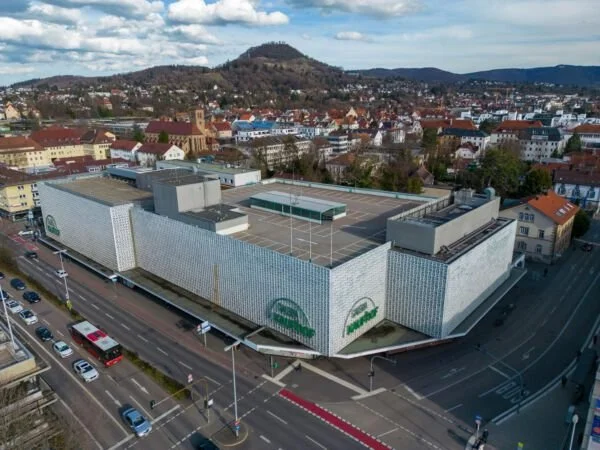 Luftaufnahme eines großen Einkaufszentrums in einer Stadt mit historischen Gebäuden und Hügel im Hintergrund. Das Einkaufszentrum hat eine moderne, weiße Fassade mit grünen Logos.