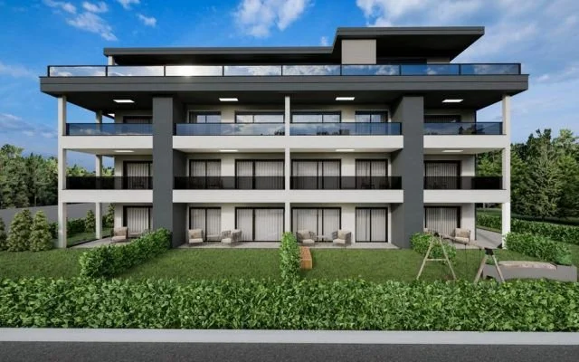 Moderne mehrstöckige Apartmentanlage mit Balkonen, Terrasse und Garten im Vordergrund, umgeben von Bäumen, unter blauem Himmel.