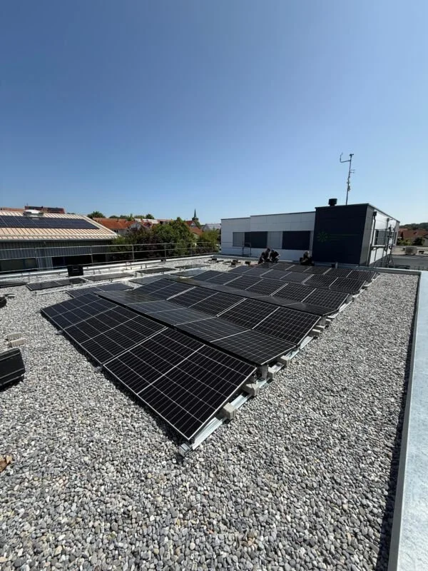 Dach mit Solarzellen auf einem Flachdach, blaue Himmel, Gebäude und Bäume im Hintergrund.