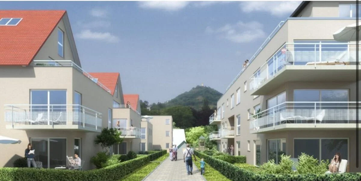 Moderne Mehrfamilienhäuser mit Balkonen und grünen Gärten, ein Fußweg zwischen den Gebäuden, im Hintergrund ein Hügel mit einer kleinen Burg