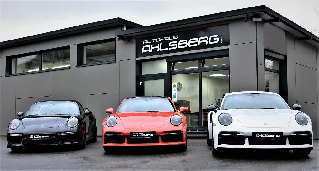 Drei Porsche-Sportwagen, schwarz, rot, weiß, stehen vor einem Autohaus namens 'Autohaus Ahlsberg' in Deutschland.