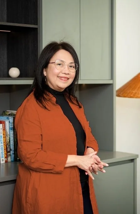 Mary Vaz, senior HR Leader and certified strategic transition coach for senior leaders, smiling and wearing an orange jacket over a black top, standing in front of a bookshelf and a gray cabinet.