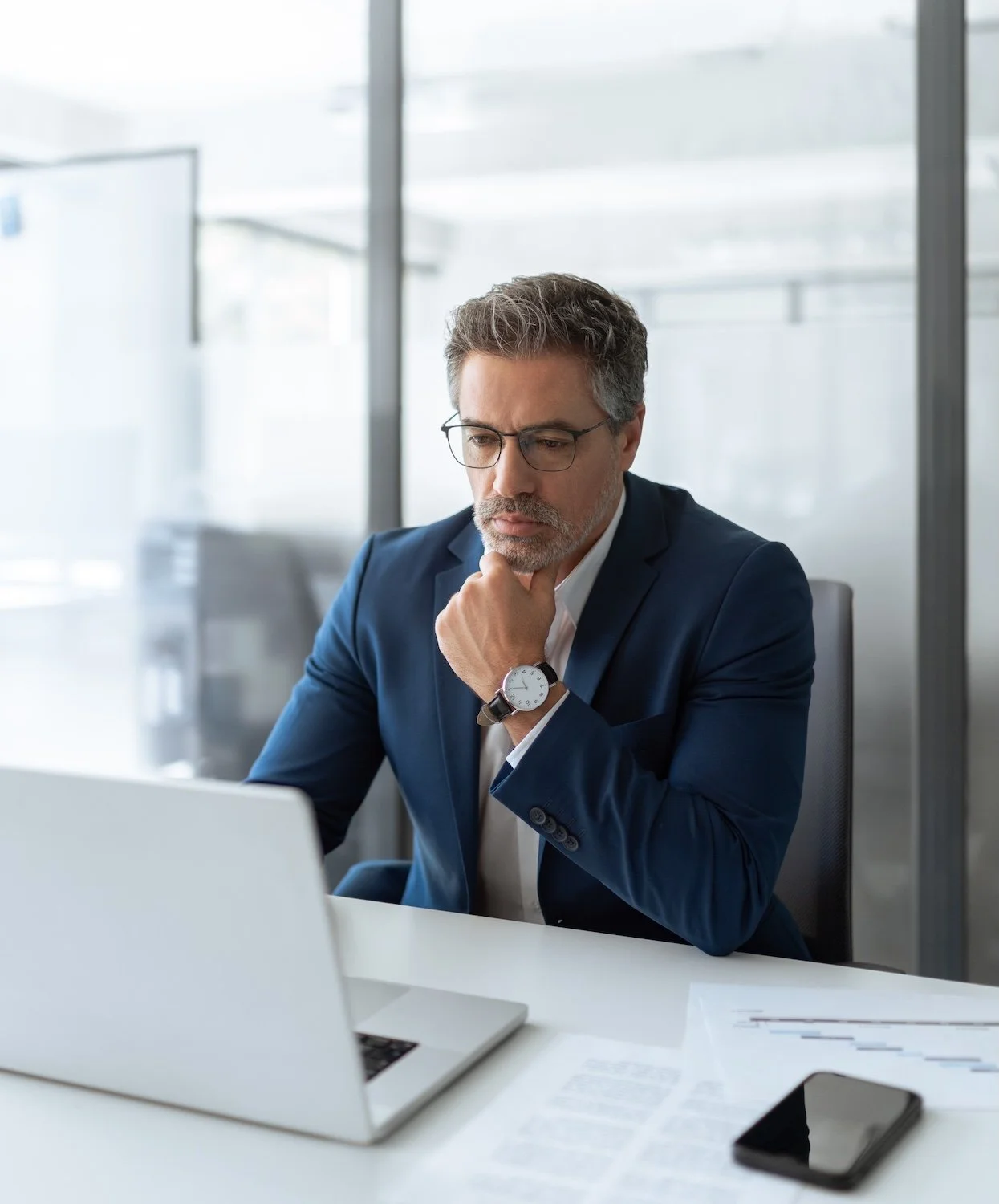 Male senior executive leader in a blue suit and glasses sitting at a white desk in an office, looking at a laptop with a thoughtful expression, with papers and a smartphone on the desk.
