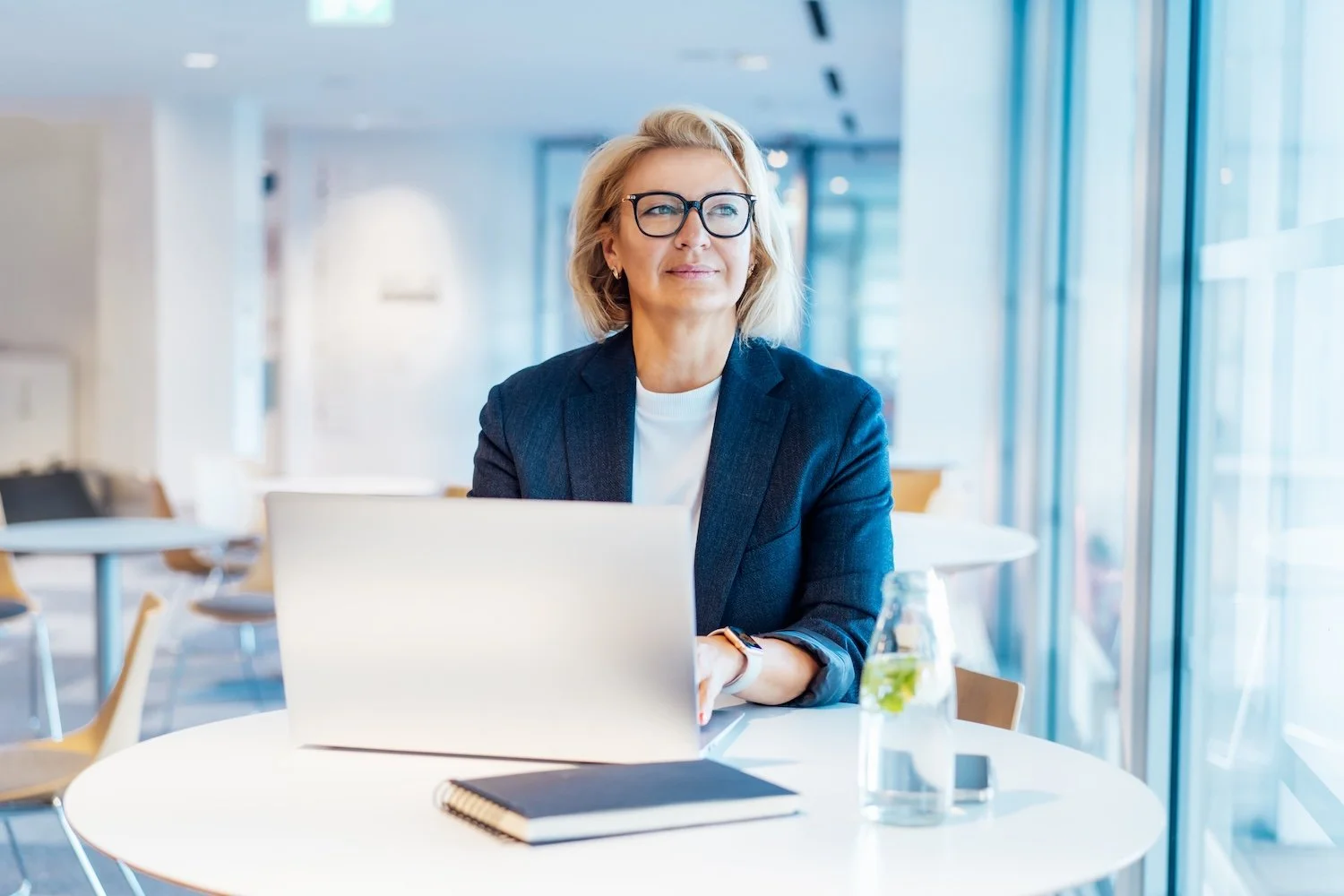 A confident middle-aged female senior leader with blonde hair, glasses, and a navy blazer working on a laptop at a round white table in a modern office or café with large windows and sunlight, looking slightly contemplative towards the left.