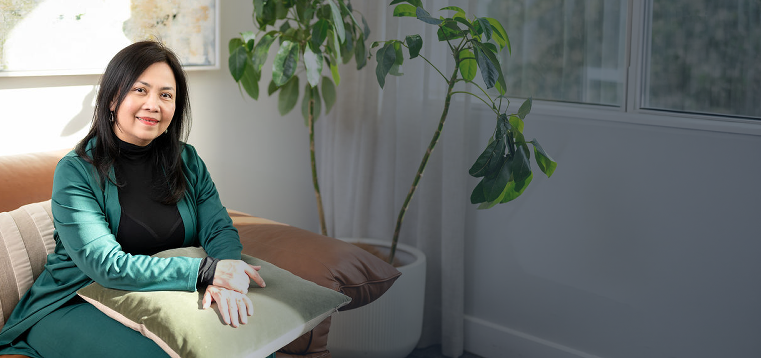 Mary Vaz, strategic transition coach for senior executive leaders, sitting on a beige sofa with a green pillow, wearing a black top and a teal blazer, inside a room with large windows and a potted plant.
