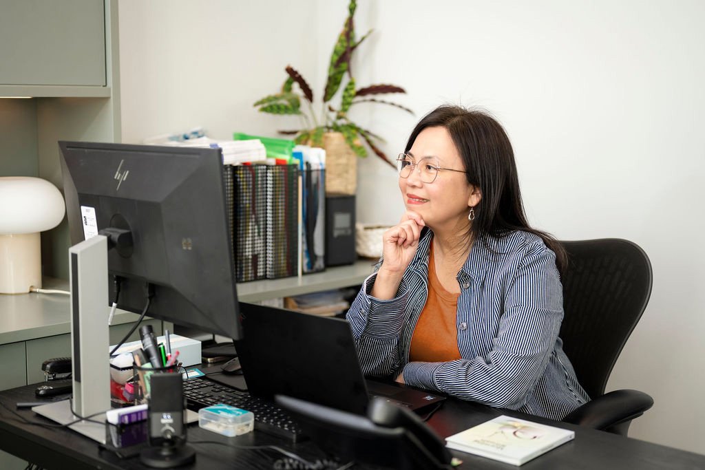 Mary Vaz, career transition coach and HR leader, sitting at a desk with a computer monitor, having a call with her client, surrounded by office supplies and a potted plant.