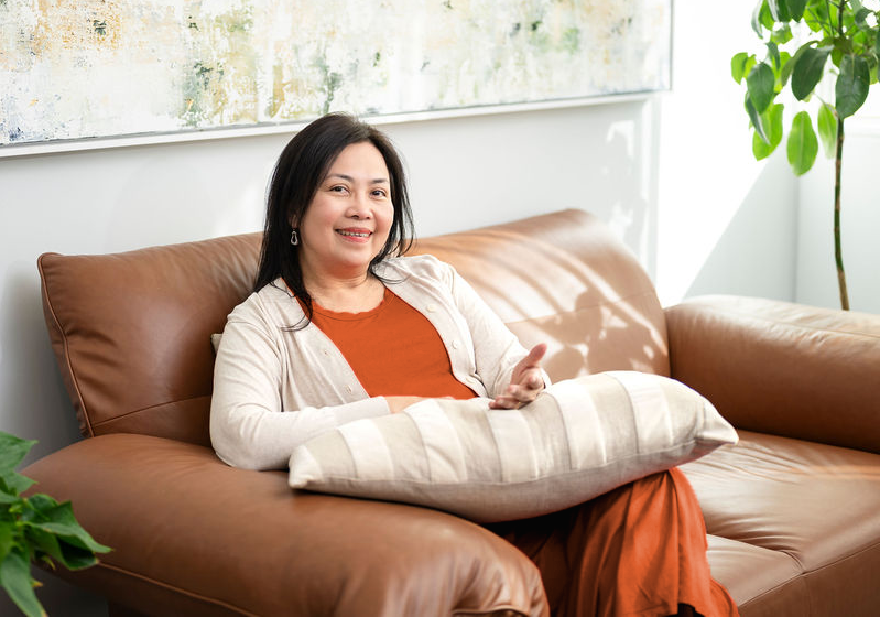 Mary Vaz, career transition coach in Australia, sitting comfortably on a brown leather sofa with a pillow in a sunlit living room, next to a green houseplant.
