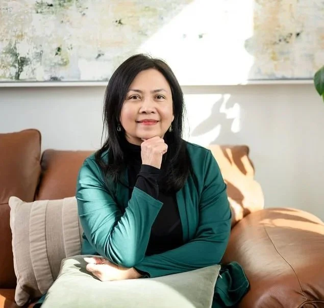 Mary Vaz, strategic transition coach for senior leaders, smiling while sitting on a beige couch in a living room, wearing a teal jacket and a black turtleneck.