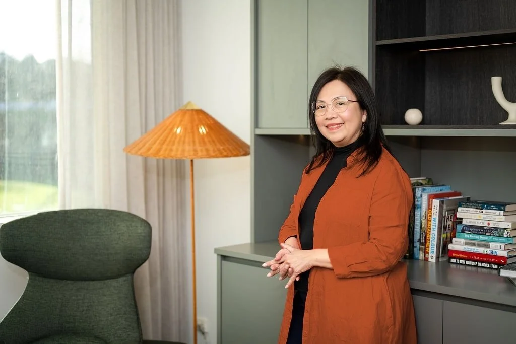 Mary Vaz, senior HR Leader and certified strategic transition coach for senior leaders, smiling and wearing an orange jacket over a black top, standing in front of a bookshelf and a gray cabinet.
