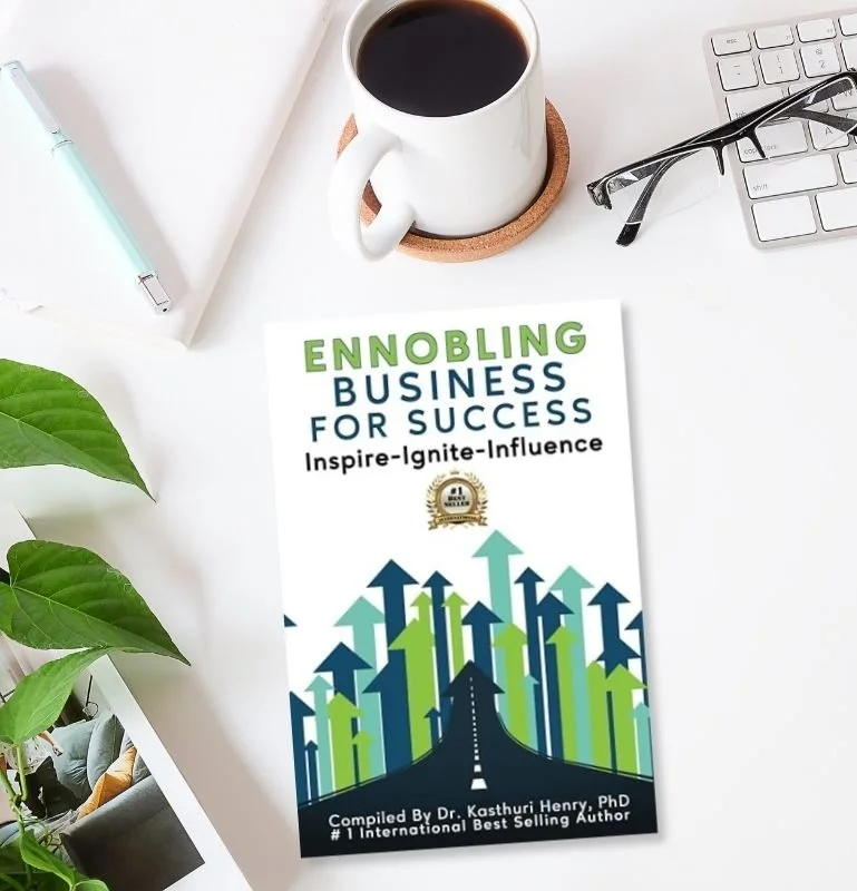 Book titled 'Ennobling Business for Success', by Dr. Kasthuri Henry, PhD, on a white desk with a coffee mug, eyeglasses, a keyboard, a pen, and some green plants.