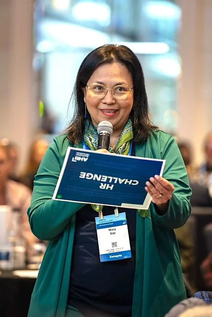 Mary Vaz, senior HR Leader and certified coach, holding a microphone at a HR conference.