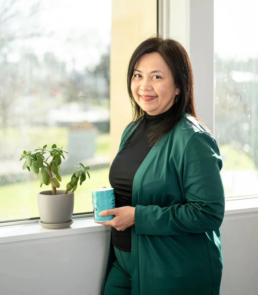 Mary Vaz, career transition coach for senior leaders, smiling, standing by a window, holding a blue mug, next to a potted plant in her office in Melbourne, Australia.