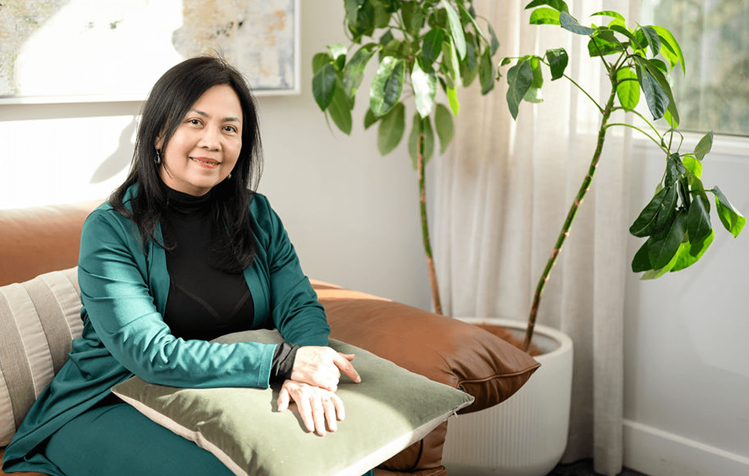 Mary Vaz, strategic transition coach for senior executive leaders, sitting on a beige sofa with a green pillow, wearing a black top and a teal blazer, inside a room with large windows and a potted plant.