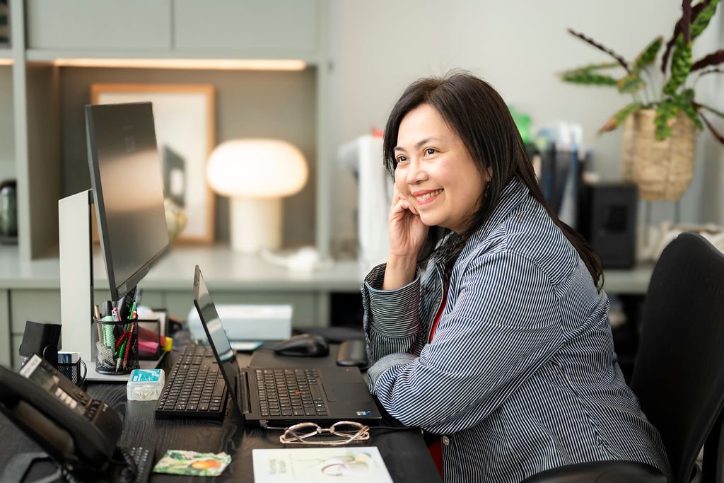 Mary Vaz, strategic transition coach, sitting at a desk in an office, smiling and looking at a computer monitor with her hand resting on the side of her face.